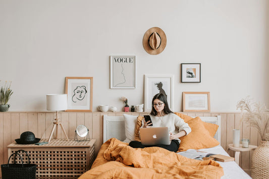 Woman sits on bed using laptop and smartphone in stylish, cozy bedroom with modern decor.