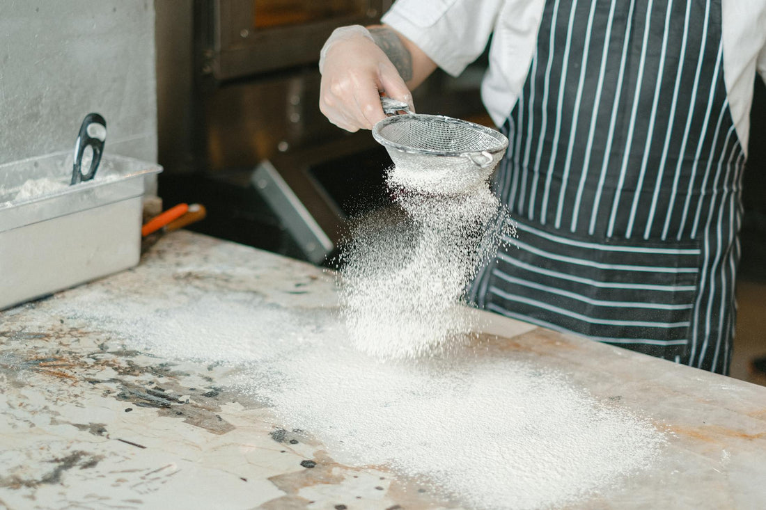 A chef in a striped apron sifting flour onto a marble countertop in a professional kitchen.